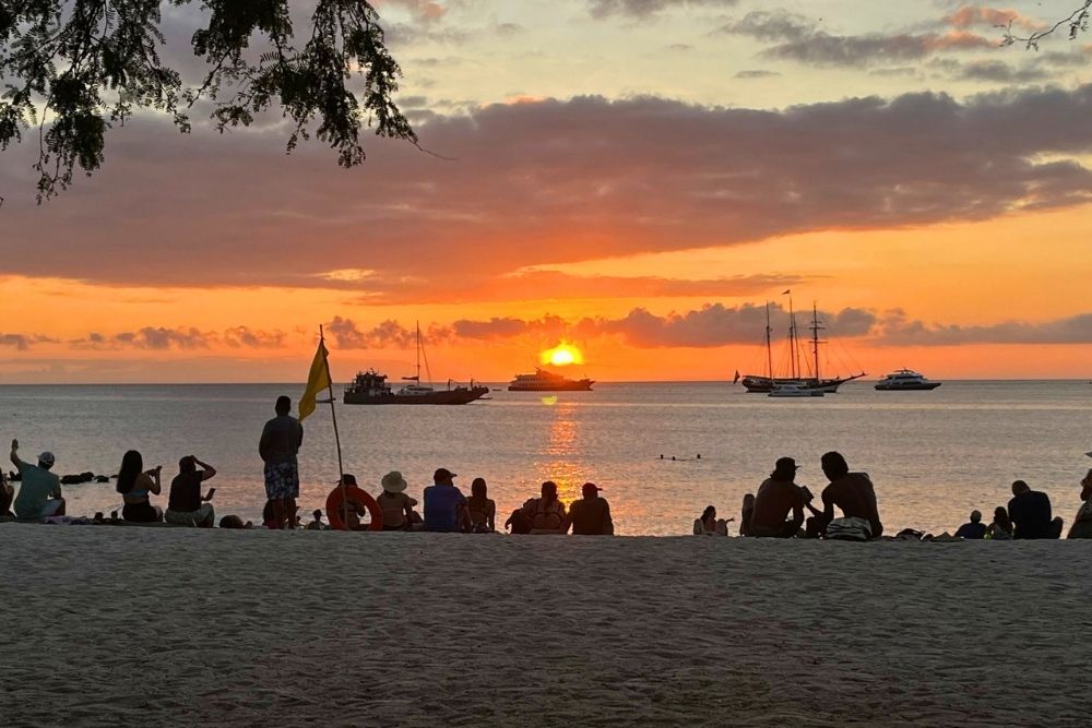People silhouetted on the sand watching the sun set over ocean vessels at Playa Mann on an 8 day Galapagos itinerary
