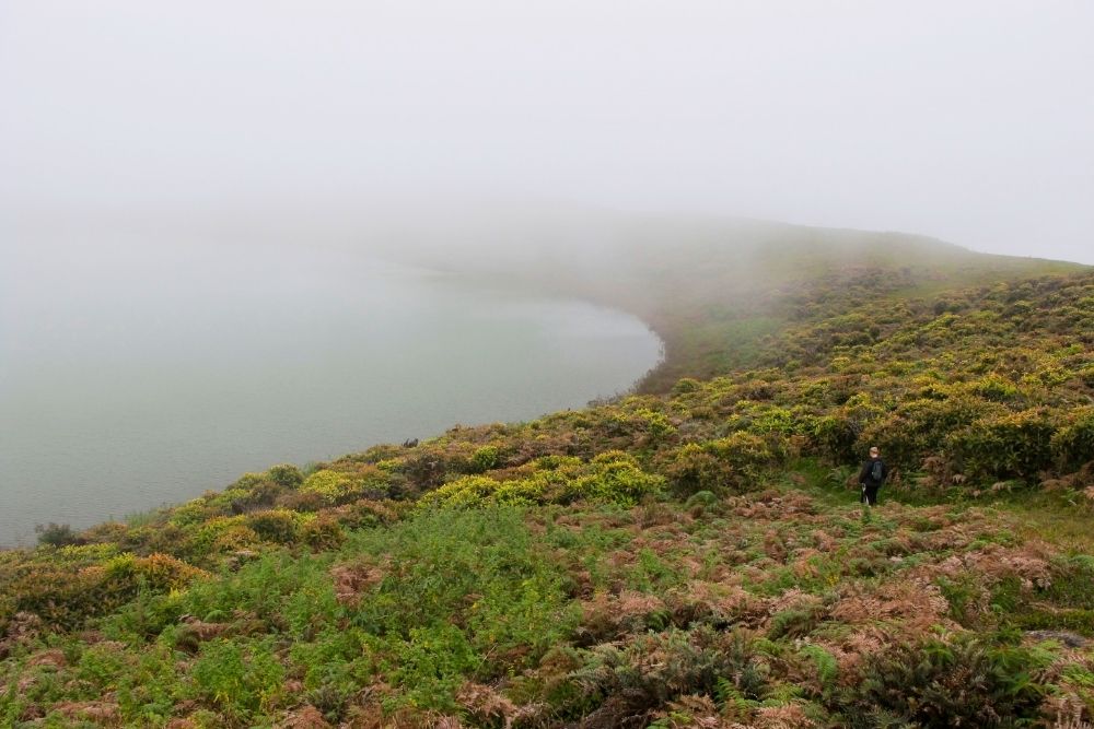 A hiker walking along lush grassy ridges surrounding foggy freshwater lake El Junco Lagoon on San Cristobal Island