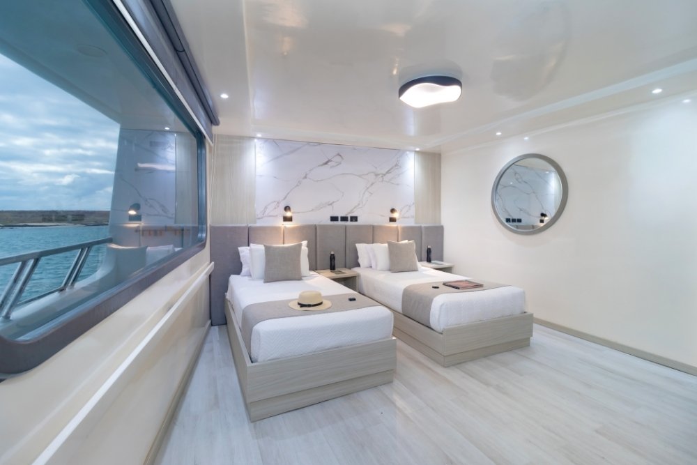 A bright and spacious twin cabin stateroom on the Galapagos Horizon vessel featuring two comfortable single beds, minimalist white decor, and a massive panoramic window with an ocean view.