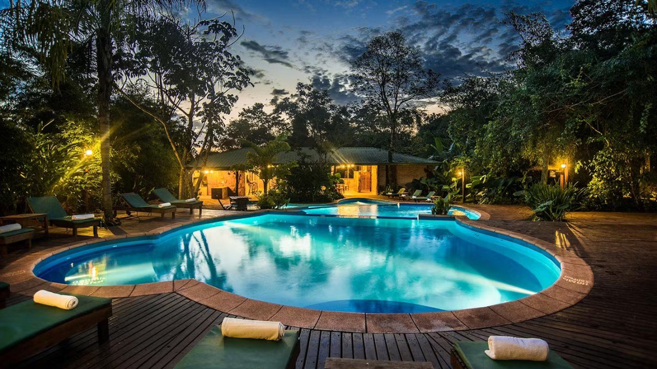 Illuminated outdoor swimming pool surrounded by lush jungle trees at twilight at La Aldea de la Selva in Puerto Iguazú