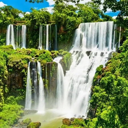 Argentina-Iguazu-Falls Rebecca Adventure Travel A vibrant view of multiple levels of powerful waterfalls plunging into a misty basin, surrounded by dense, bright green jungle foliage during an Iguazu excursion.