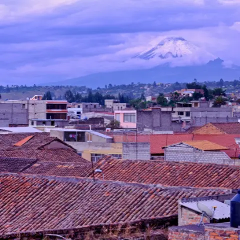 Latacunga Rebecca Adventure Travel View of Latacunga city rooftops with the majestic snow-capped Cotopaxi volcano towering in the background under a cloudy sky.