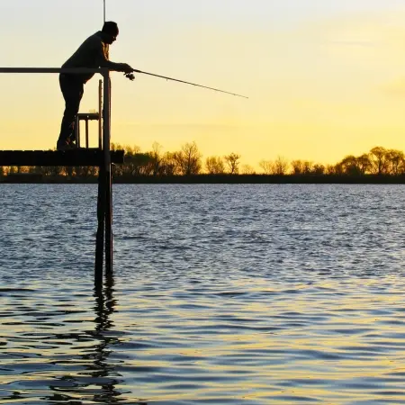Parana-River Rebecca Adventure Travel The silhouette of a man fishing from a wooden pier at sunset, with golden light reflecting off the ripples of the water during a Parana Luxury adventure.