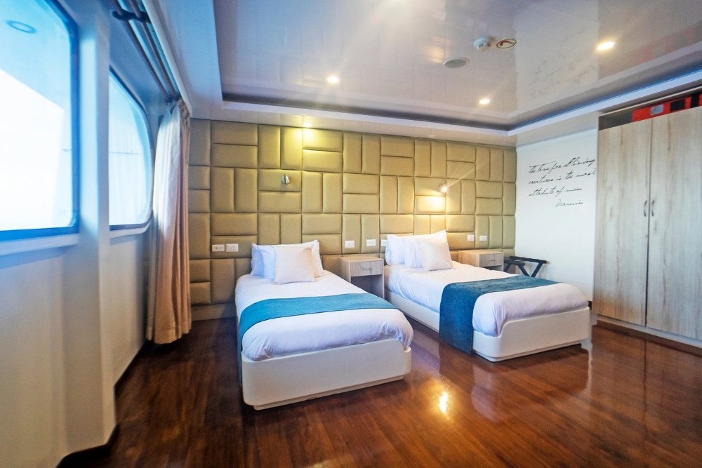 A bright and elegant twin cabin on the Petrel Luxury Cruise featuring two separate beds with blue accents, polished wooden floors, a large wardrobe, and wide windows with ocean views.