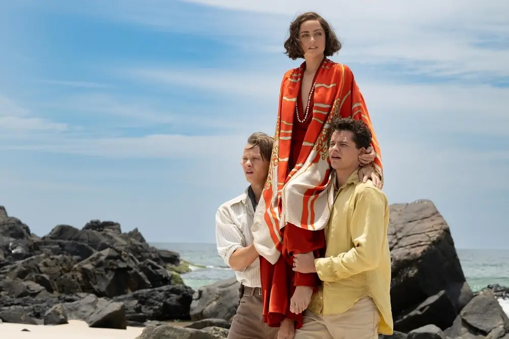 two men carry a woman in a red dress across a rocky beach in a scene from eden the movie.
