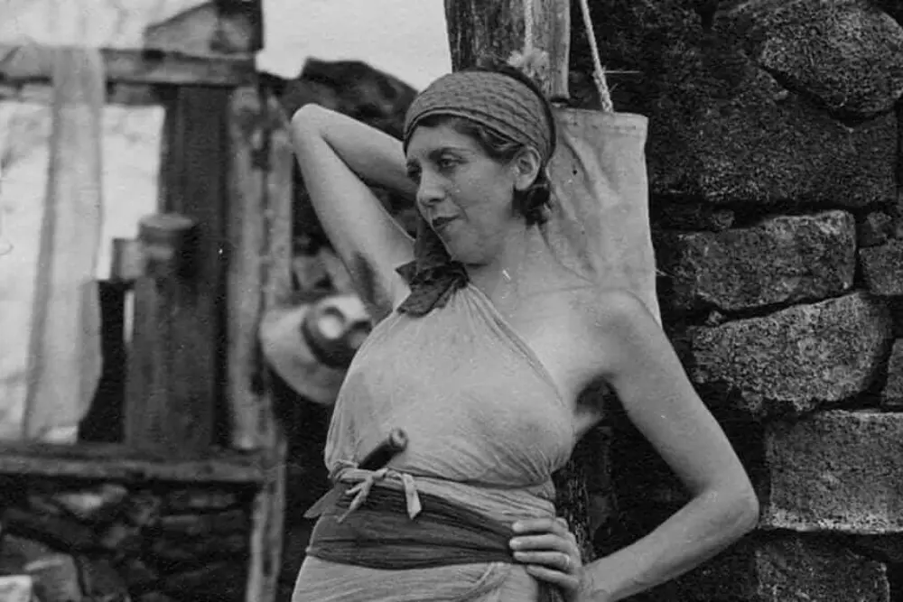 a black and white photo of the baroness wearing a headscarf and a draped outfit while leaning against a stone wall.