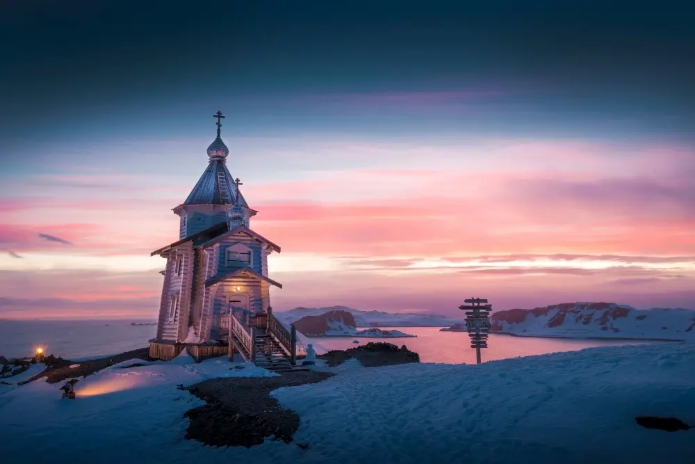 a small, blue wooden church with a silver spire stands illuminated against a vibrant pink and purple sunset on a snow-covered hill at the russian station of the antarctica.