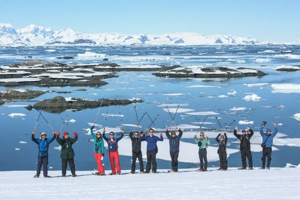 a group of travelers stands atop a pristine snow-covered slope, raising their trekking poles in celebration against a backdrop of glassy blue water and floating icebergs during a session of snowshoeing in antarctica.