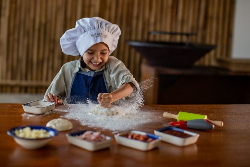 child wearing a chef hat at galapagos safari camp enjoying a hands on cooking class, preparing dough at a wooden table with fresh ingredients